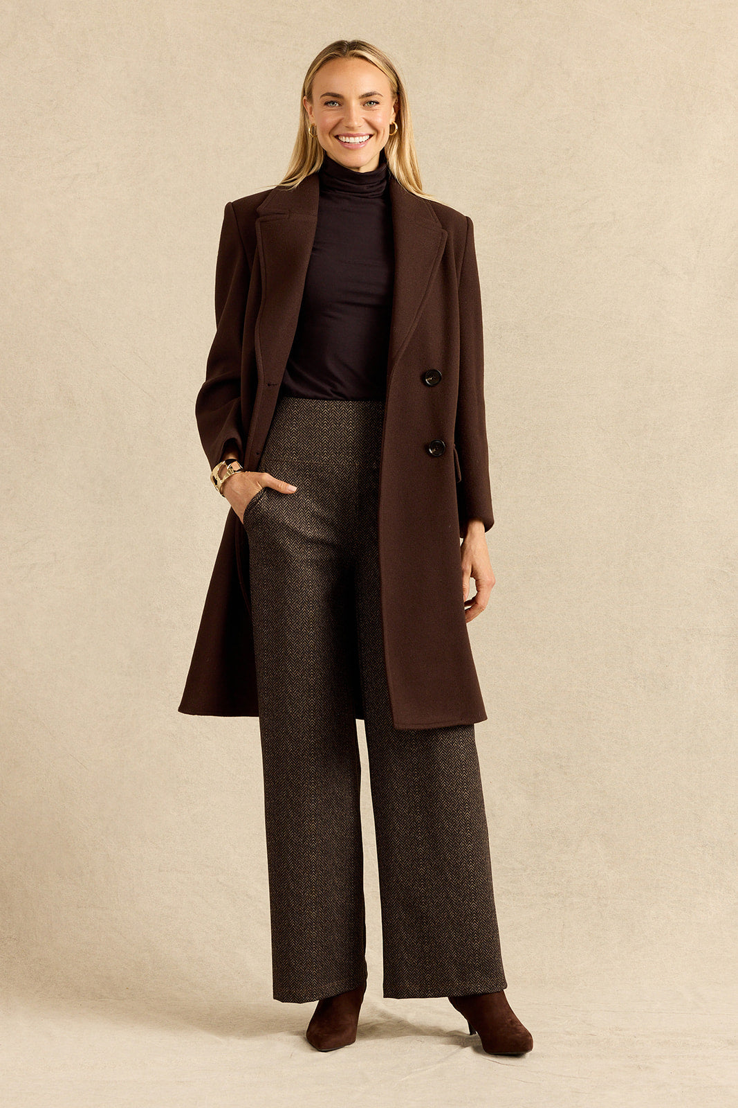 A woman stands against a warm beige studio backdrop wearing a long dark brown double-breasted coat over a dark turtleneck, brown herringbone wide-leg trousers, and brown ankle boots.