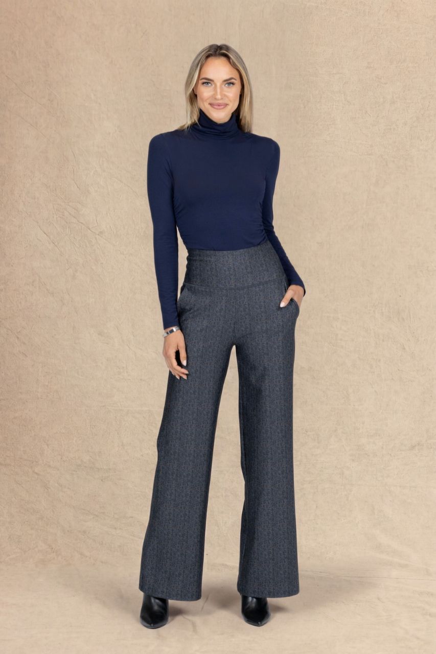 A woman modeling a navy blue turtleneck long-sleeve top and high-waisted, wide-leg charcoal gray pinstripe trousers, standing against a textured beige backdrop.