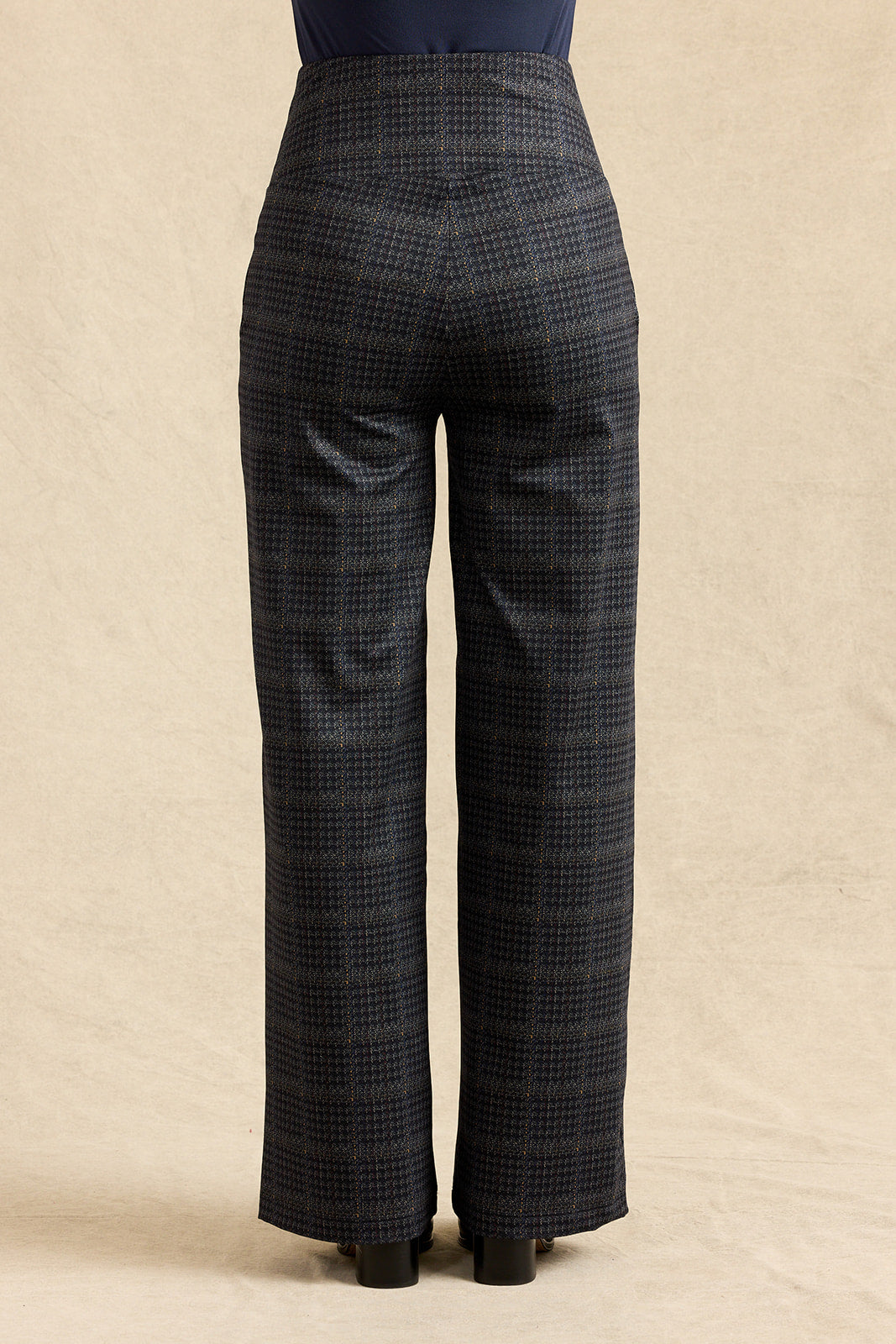 Back view of a person wearing high-waisted, wide-leg trousers in a dark gray micro-check pattern, paired with a dark top and black heels, against a neutral beige backdrop.