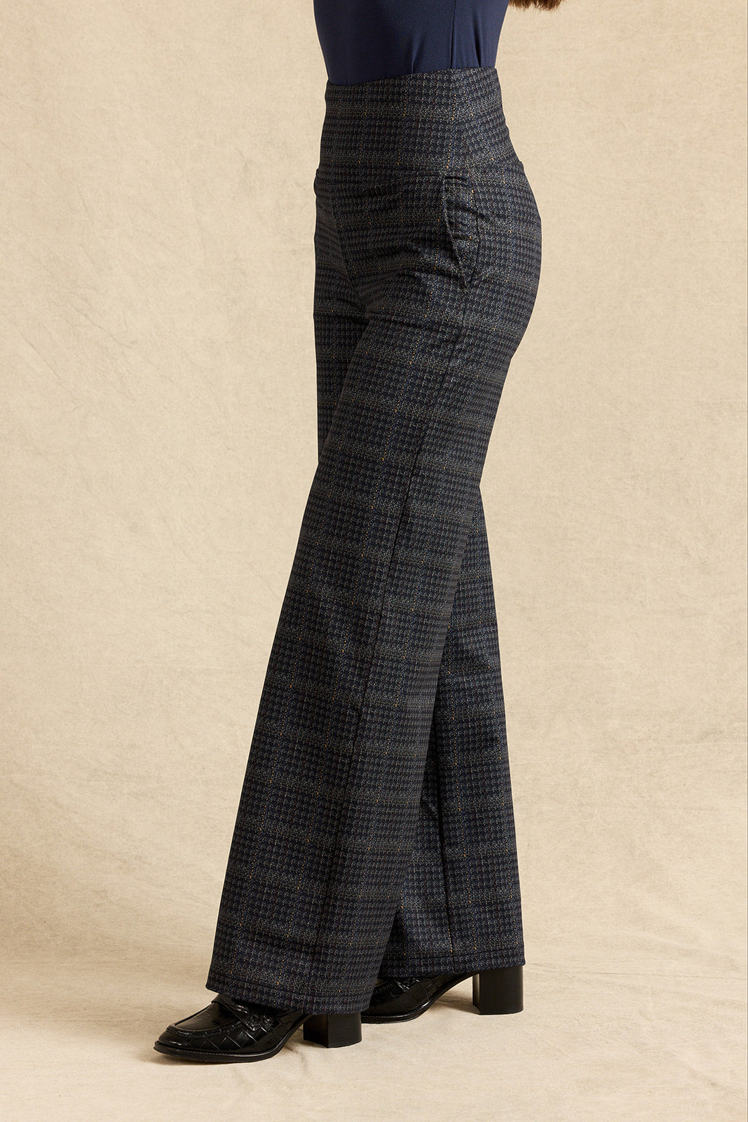 A person shown from the waist down wearing dark navy plaid wide leg trousers with a subtle windowpane pattern, paired with black shoes, against a neutral beige backdrop.