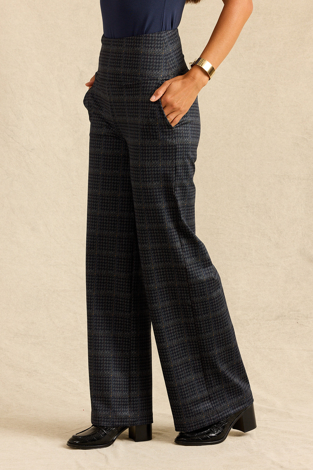 A model wearing high-waisted, wide-leg trousers in a dark grey and black micro-check plaid pattern, paired with black leather block-heel mules, against a neutral beige backdrop.