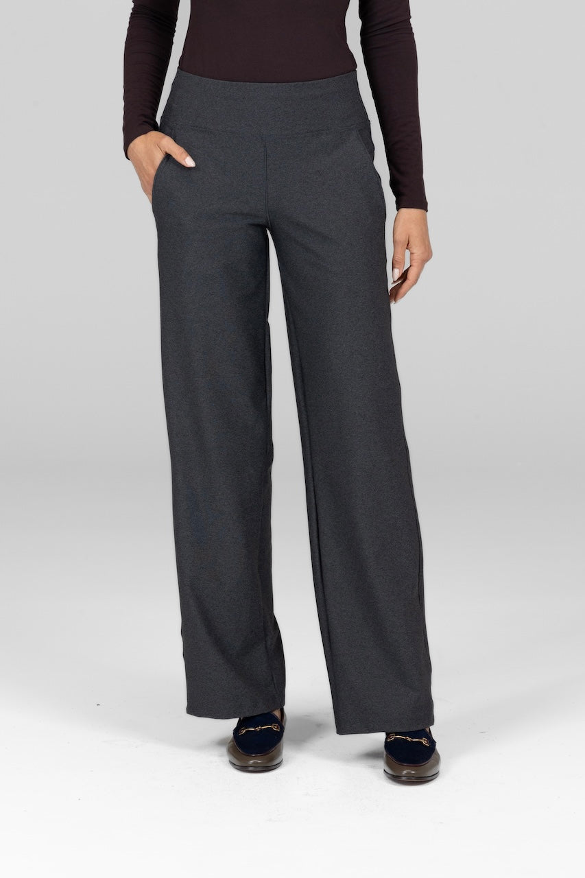 A woman standing with hands in pockets, wearing charcoal grey traveler pants with a supportive waistband.