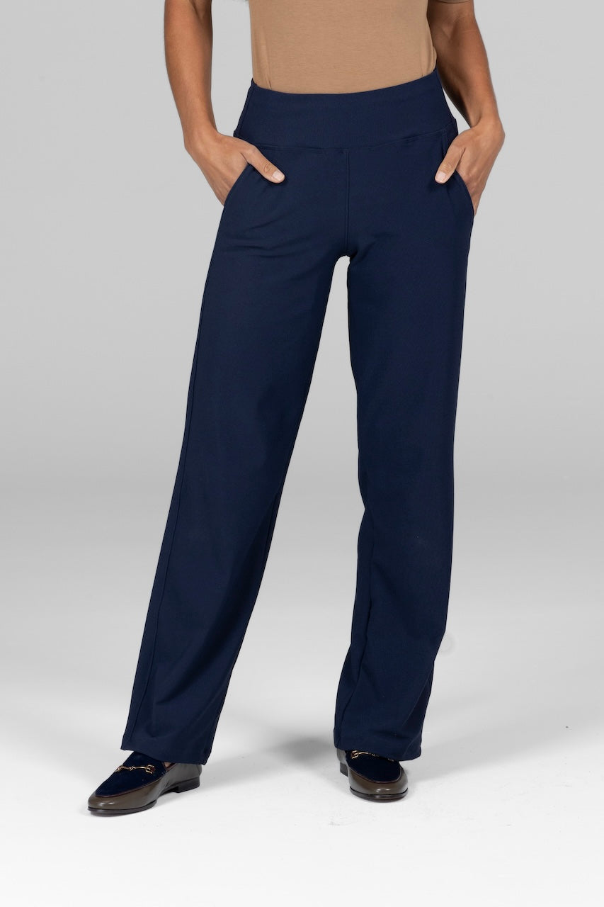 A woman standing wearing marine navy colored traveler pants with a supportive waistband and pockets.