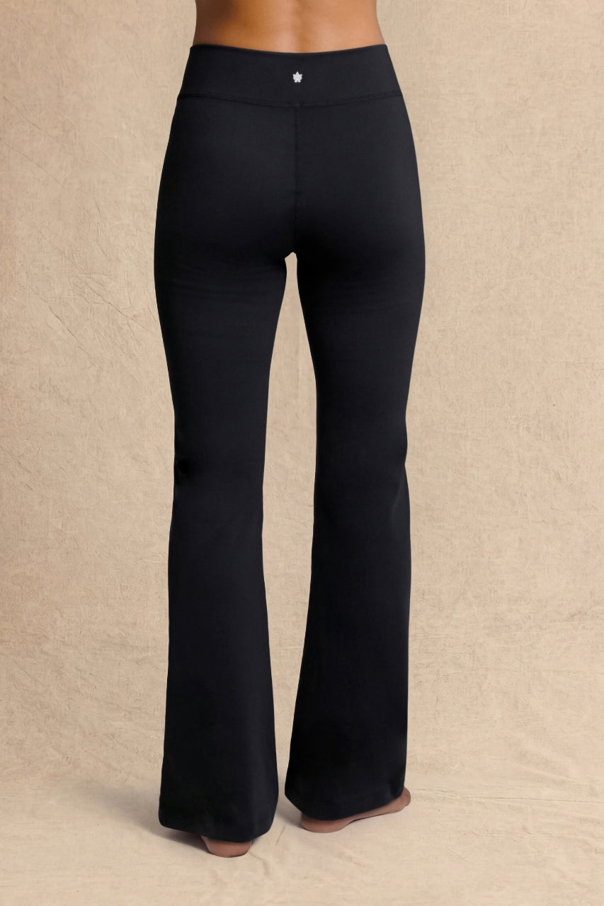 Black flared pants shown from the back on a model, featuring a mid-waisted waistband with a small silver button detail, set against a neutral beige textured backdrop.