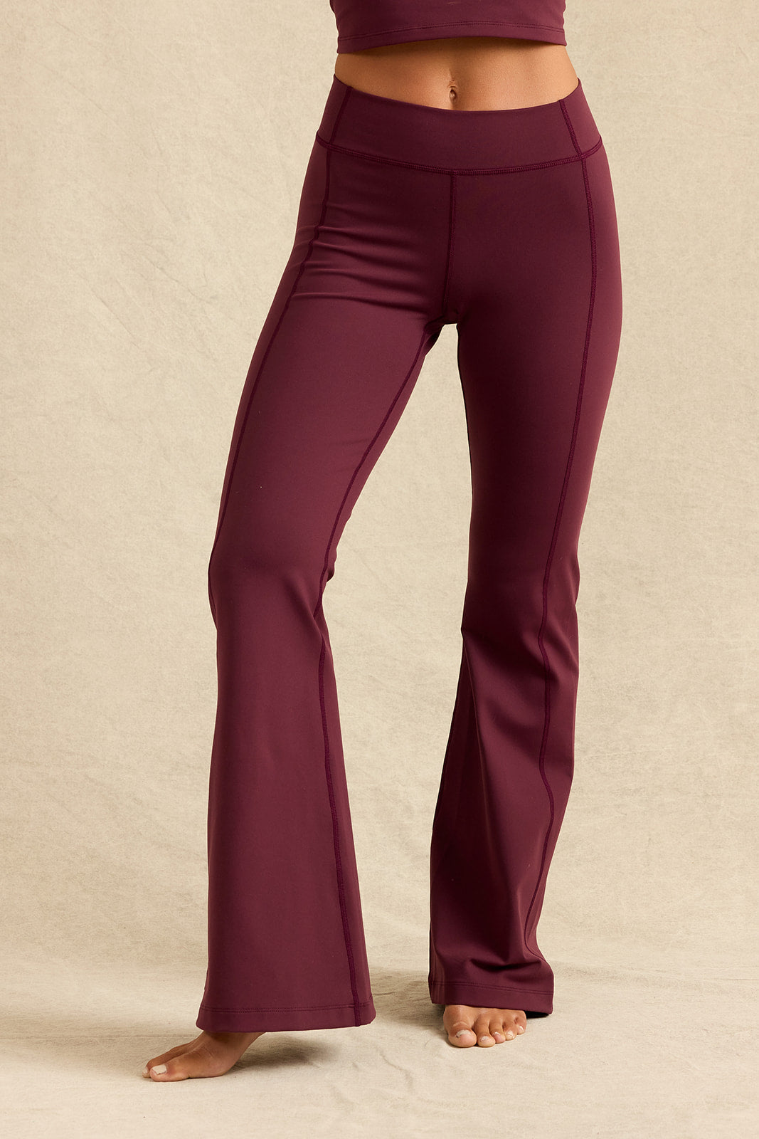 A person wearing high-waisted, wide-leg flared pants in a deep burgundy shade, standing barefoot against a neutral beige studio backdrop.