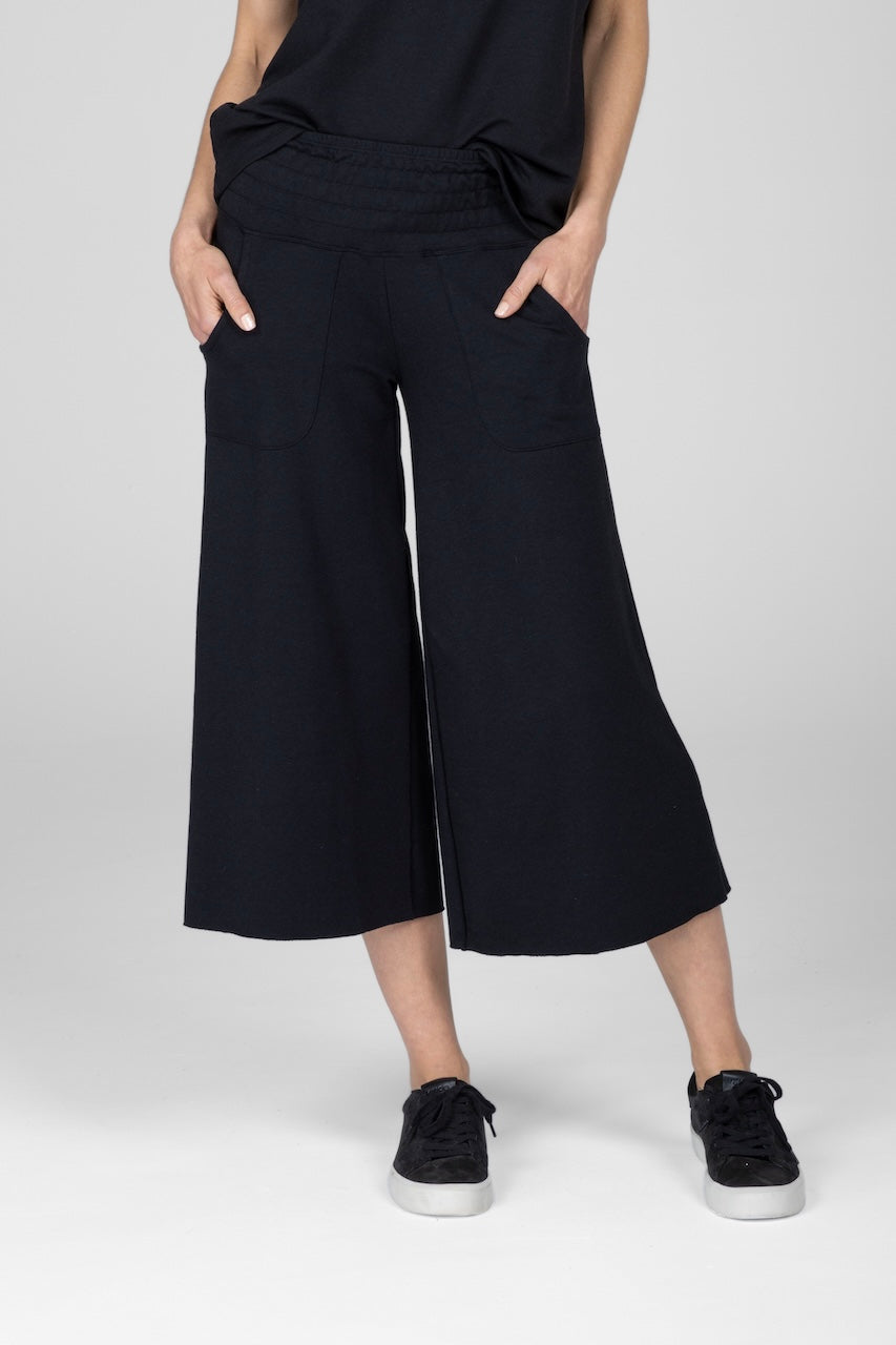 Front view of woman wearing cropped black boho yoga pants paired with a black top and black sneakers, model with hands in pockets.