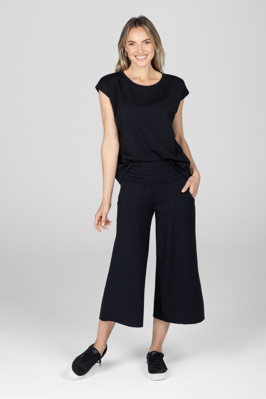 A woman standing, wearing a black french terry top with short sleeves and matching black french terry cropped lounge pants, with one hand in the front pocket and one hand at her side