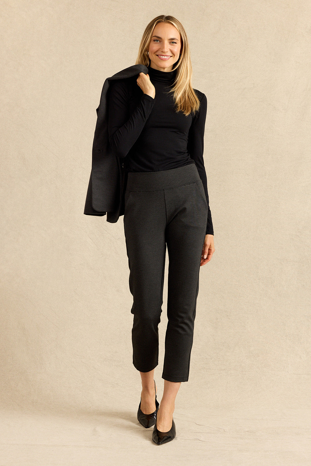 A woman modeling a black turtleneck top, charcoal grey cropped trousers, and a charcoal blazer draped over her shoulder, standing against a textured beige studio backdrop.