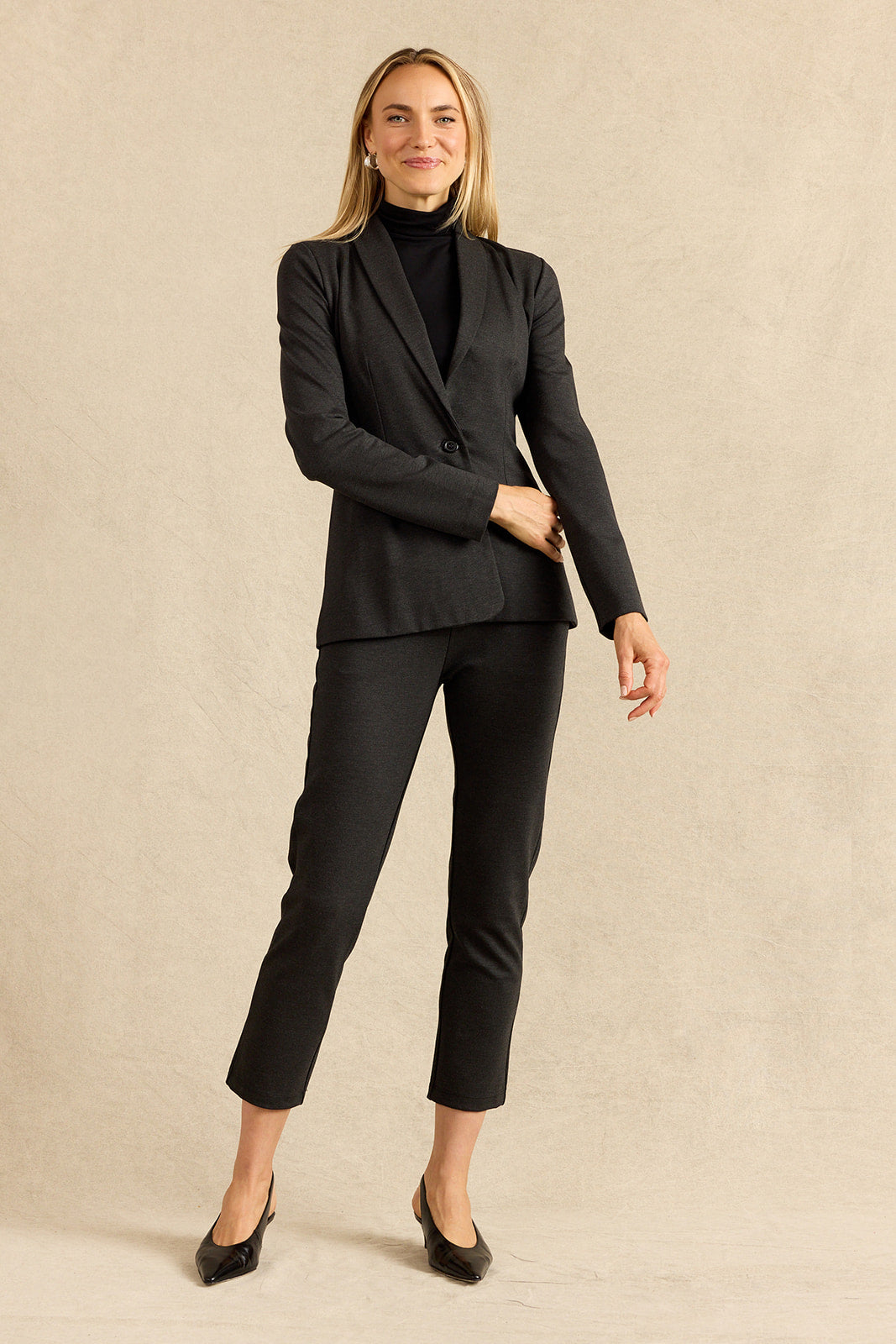 A woman modeling a charcoal tailored pantsuit consisting of a single-breasted blazer and cropped trousers, paired with a black turtleneck and black pointed heels, against a neutral beige studio backdrop.