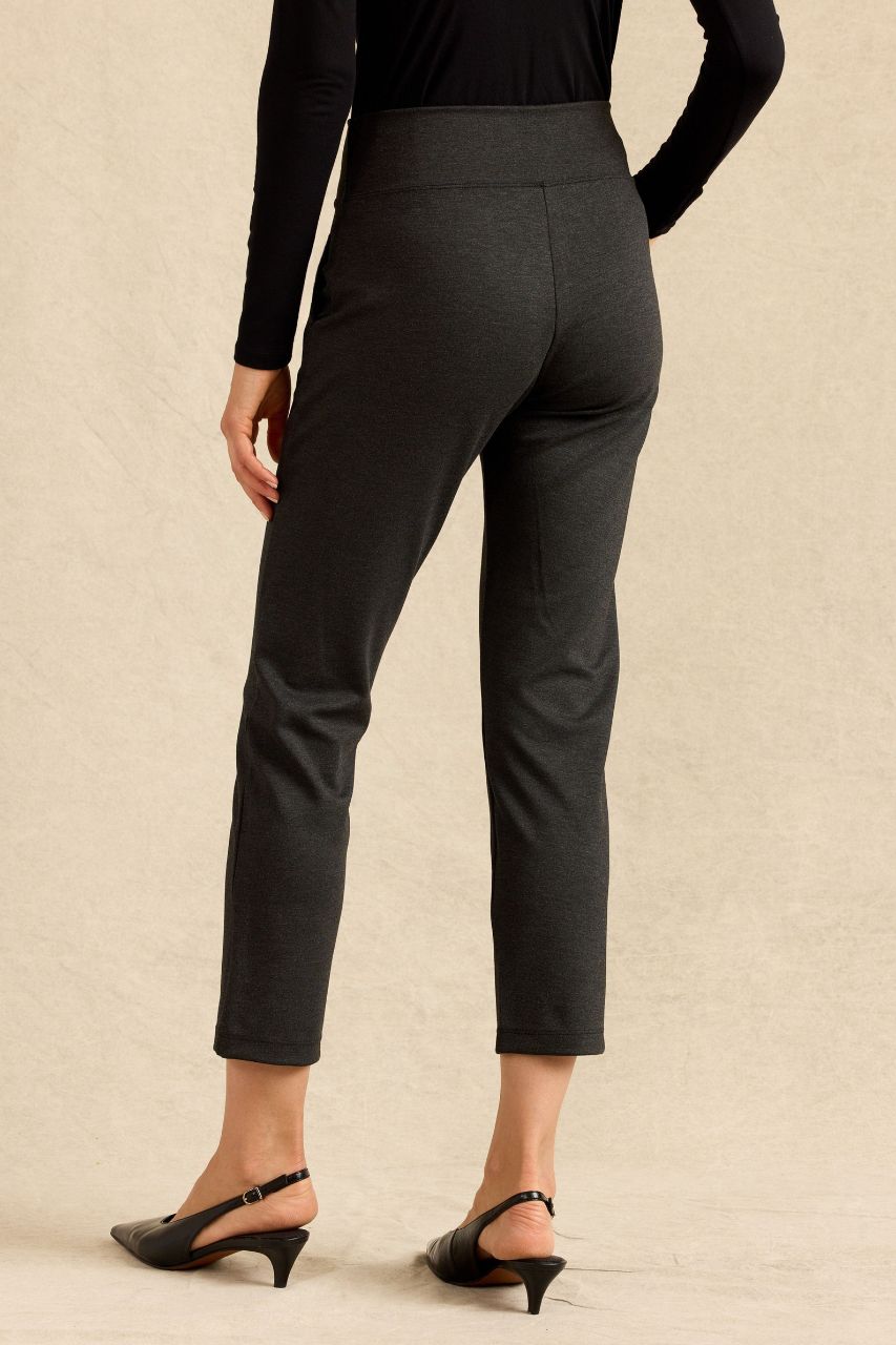 Rear view of a model wearing dark charcoal gray cropped pants with a black long-sleeve top and black slingback heels, set against a textured beige background.