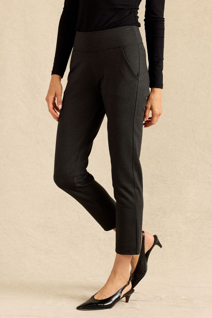 A model wearing charcoal tailored trousers with side pockets, paired with a black long-sleeve top and black slingback heels, standing against a textured beige studio backdrop.