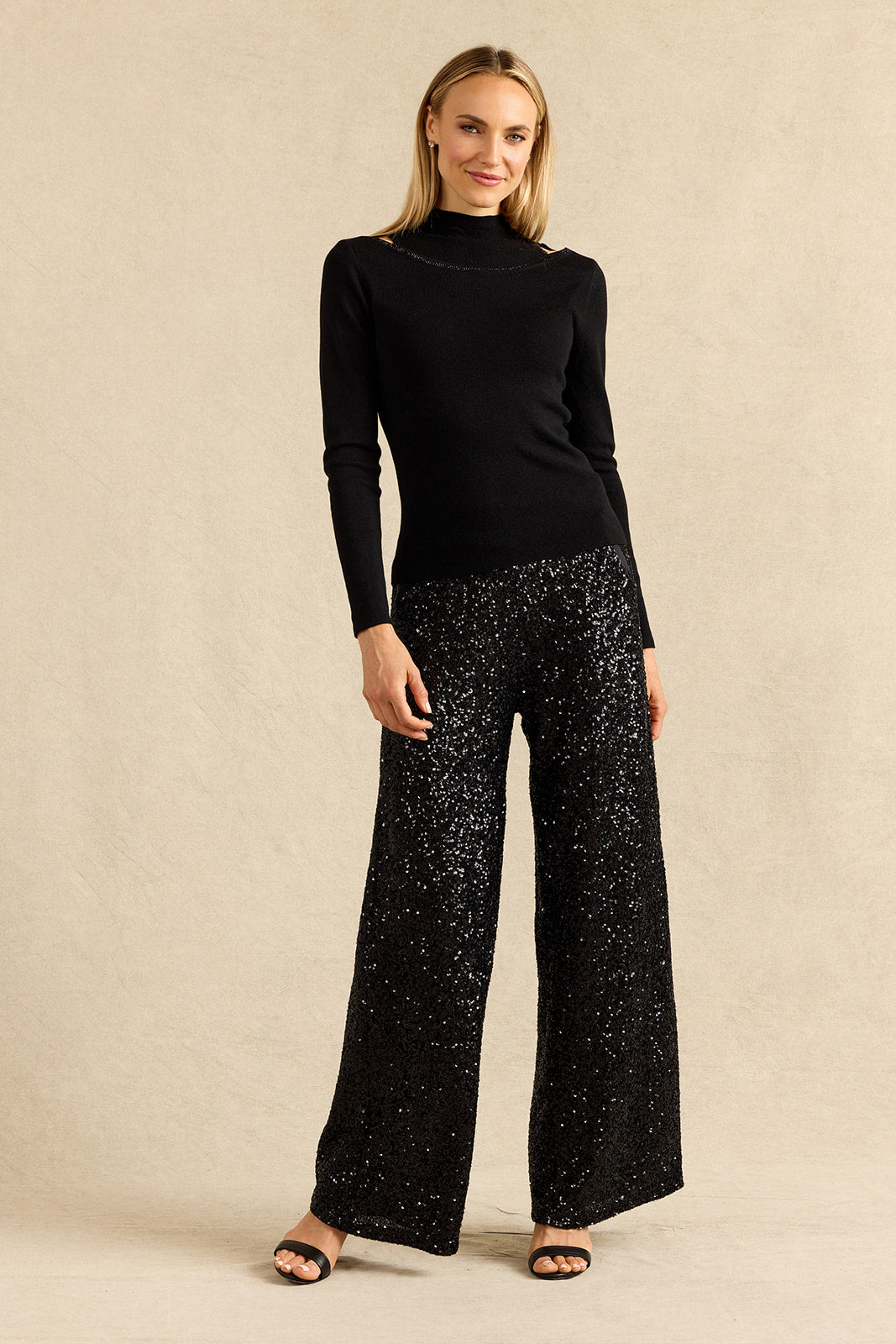A woman modeling a black long-sleeve mock neck top paired with black wide-leg pants covered in silver sequins, standing against a textured beige studio backdrop.