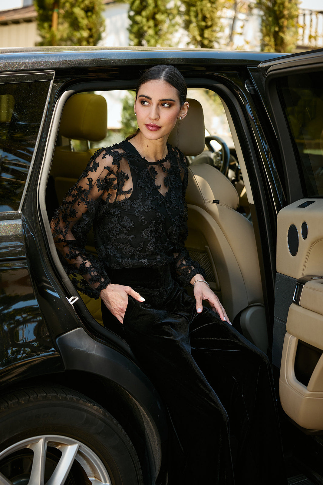 A woman in a black lace long-sleeve top and black velvet trousers sits in the open rear door of a black SUV with beige interior seats.