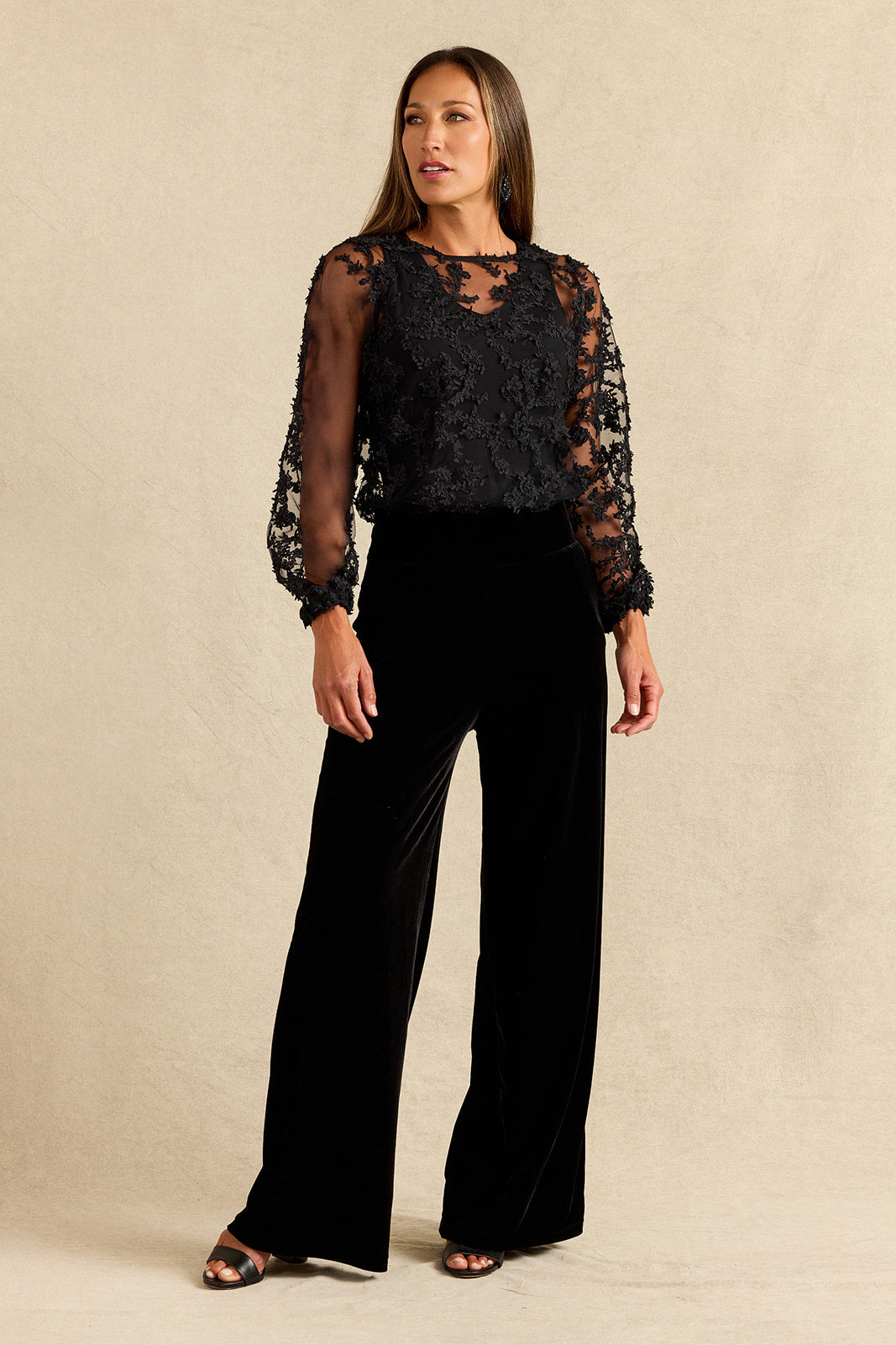A woman modeling a black lace long-sleeve top paired with black wide-leg velvet pants, standing against a neutral beige studio backdrop.