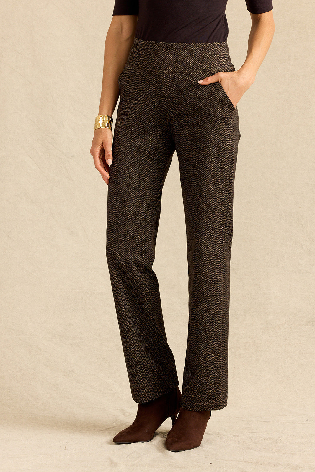 A person wearing high-waisted, straight-leg brown herringbone trousers, paired with a brown top and brown ankle boots, standing against a textured beige backdrop.
