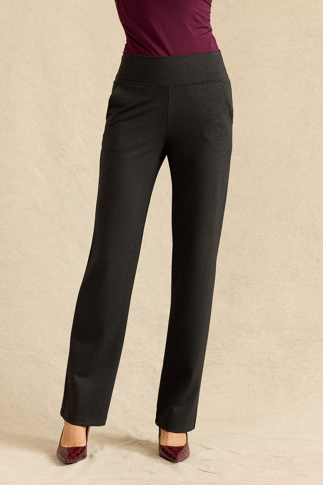 A person wearing grey straight-leg trousers with a wide waistband, paired with a burgundy top and burgundy pointed-toe heels, standing against a textured beige studio backdrop.