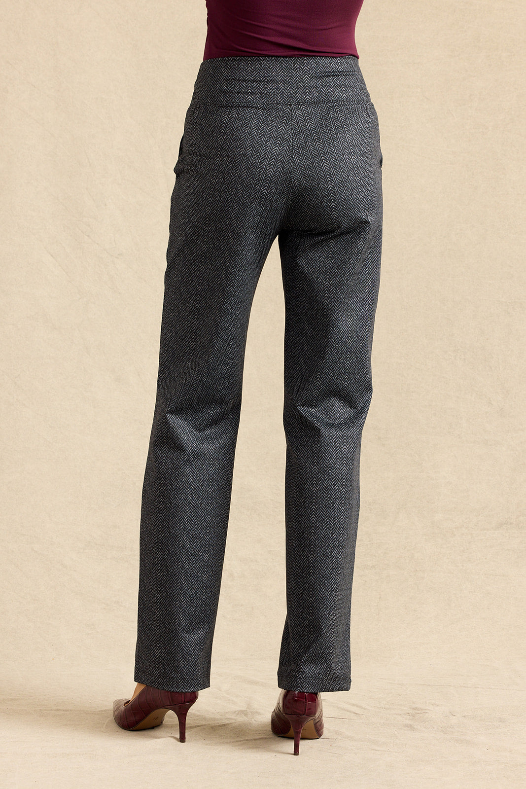 A person shown from the back wearing dark gray herringbone trousers, a burgundy top, and brown heels against a beige studio backdrop.