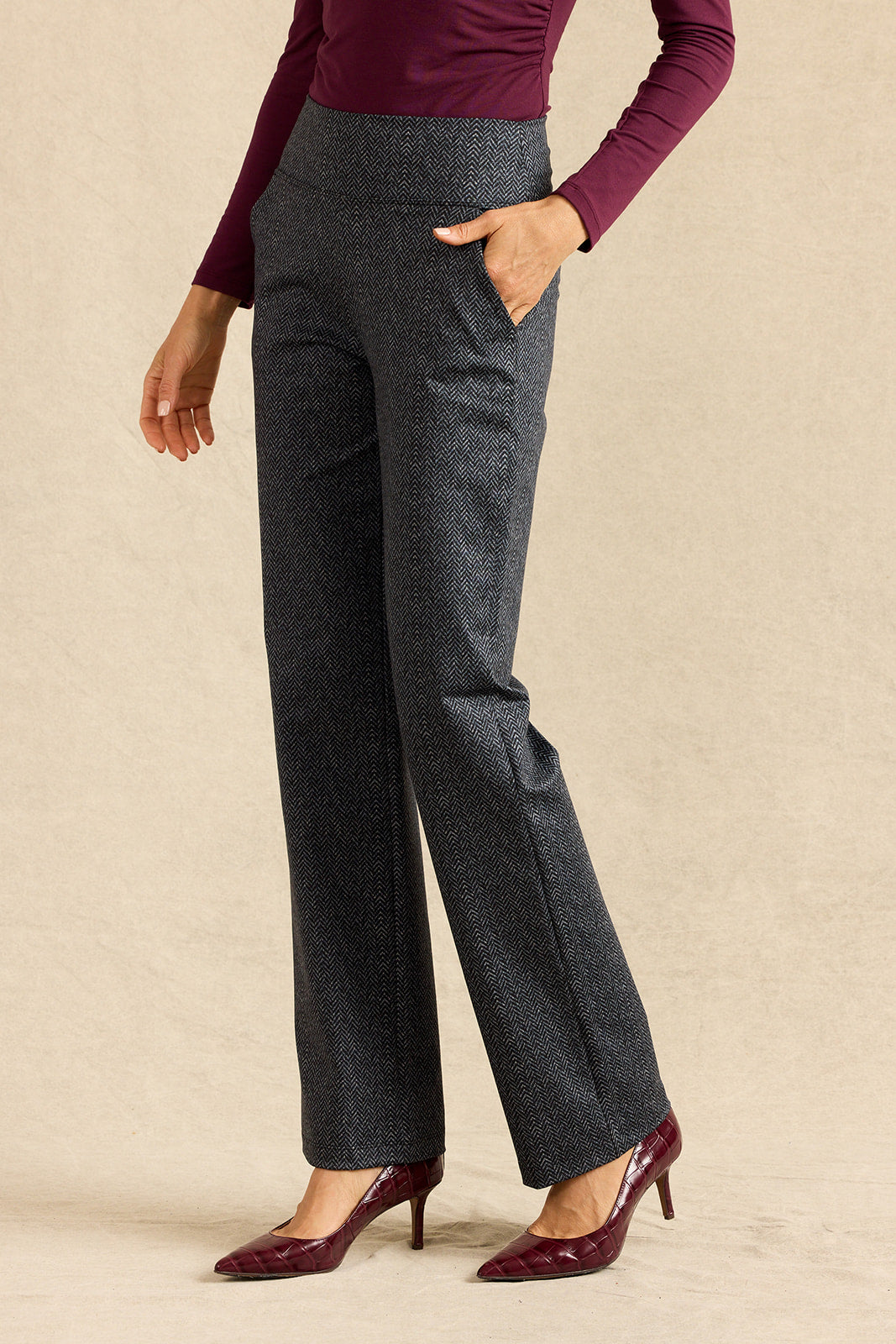 A model wearing high-waisted, straight-leg charcoal trousers with a subtle herringbone pattern, styled with a burgundy long-sleeve top and burgundy patent leather pumps against a textured beige studio backdrop.