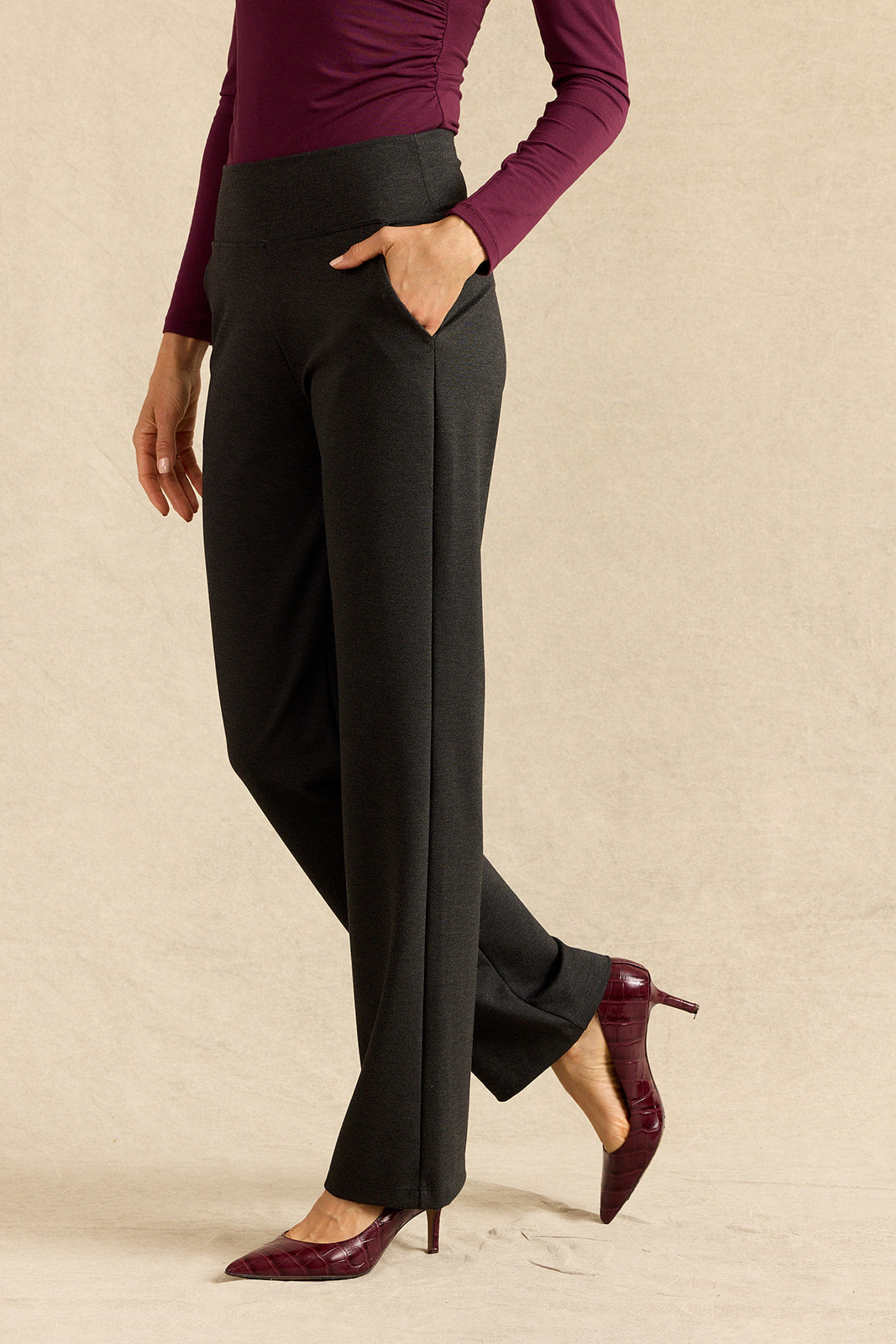 A model wearing grey wide-leg trousers with side pockets, paired with a dark burgundy long-sleeve top and matching pointed-toe heels, standing against a neutral beige backdrop.