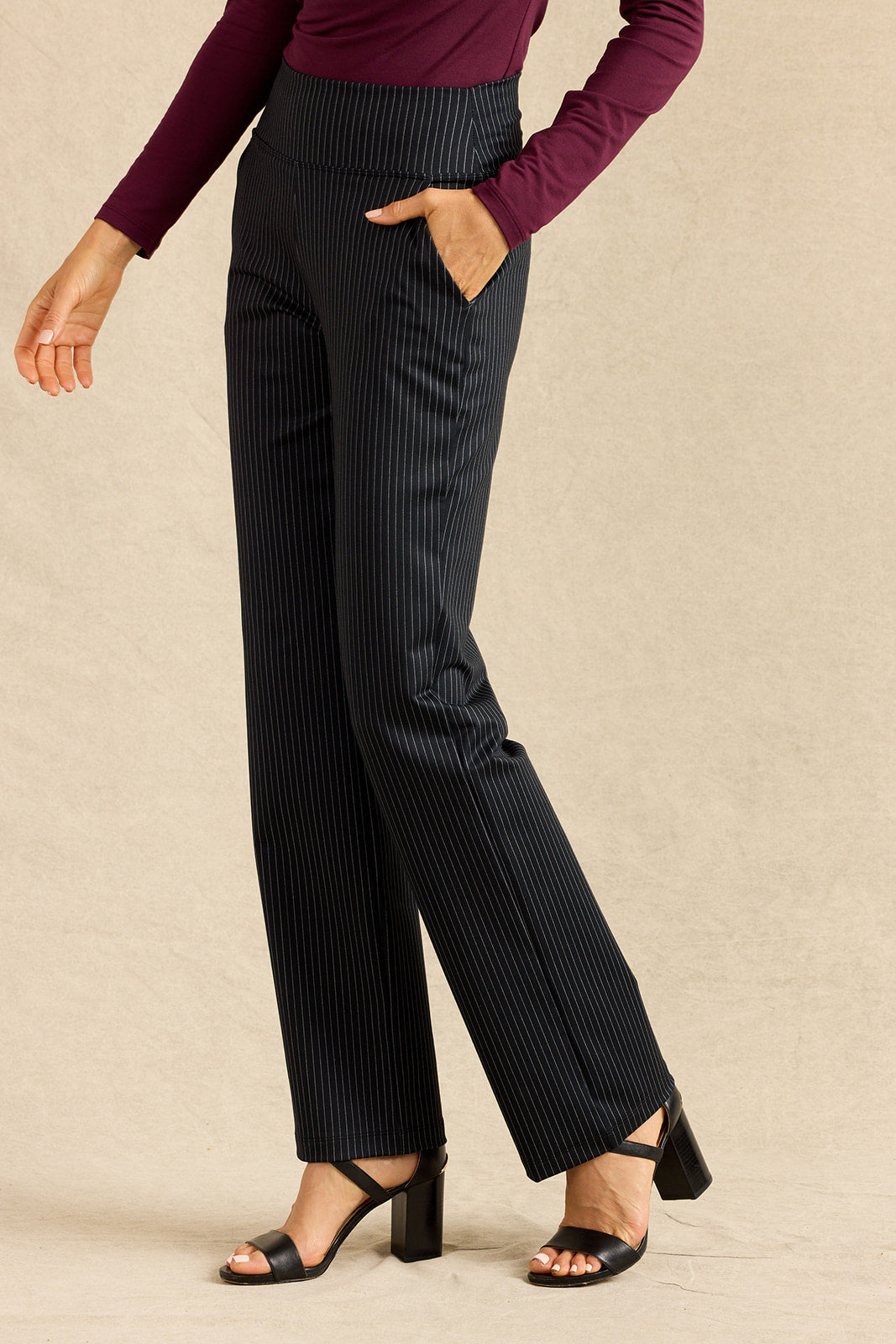 A model wearing high-waisted, wide-leg pinstripe trousers in black with thin white stripes, paired with a dark purple long-sleeve top and black block-heel sandals, set against a neutral beige studio background.
