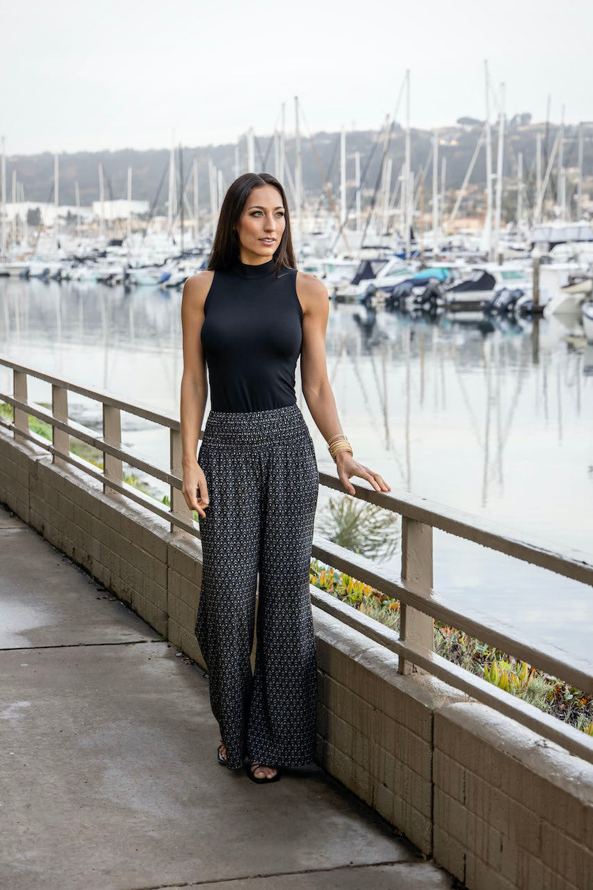 Woman by the water wearing a black top paired with the Portofino Travel Pant in woodblock print, creating a stylish and relaxed look perfect for a scenic day out