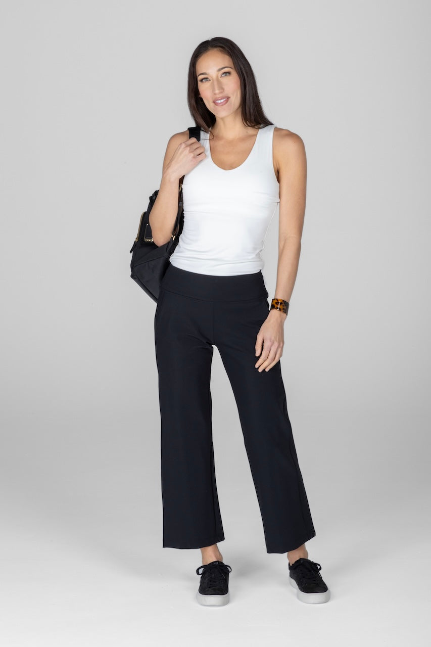 A woman standing against a white background wearing black ankle-length pants and a white sleeveless top. She has a shoulder bag and wearing black shoes.
