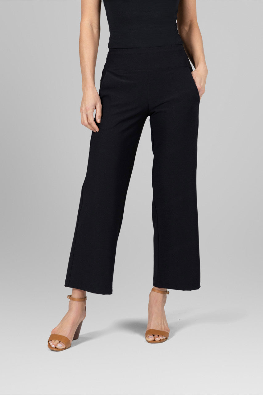 A person standing wearing black ankle pants and a black top, with tan ankle strap sandals.