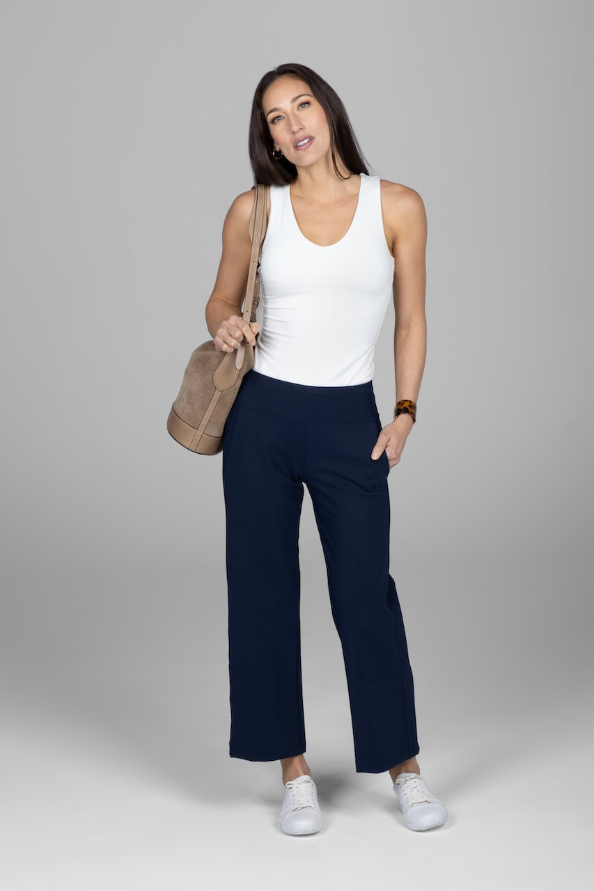A woman standing against a grey background wearing a white sleeveless top and navy blue pants, with a beige handbag and white shoes.