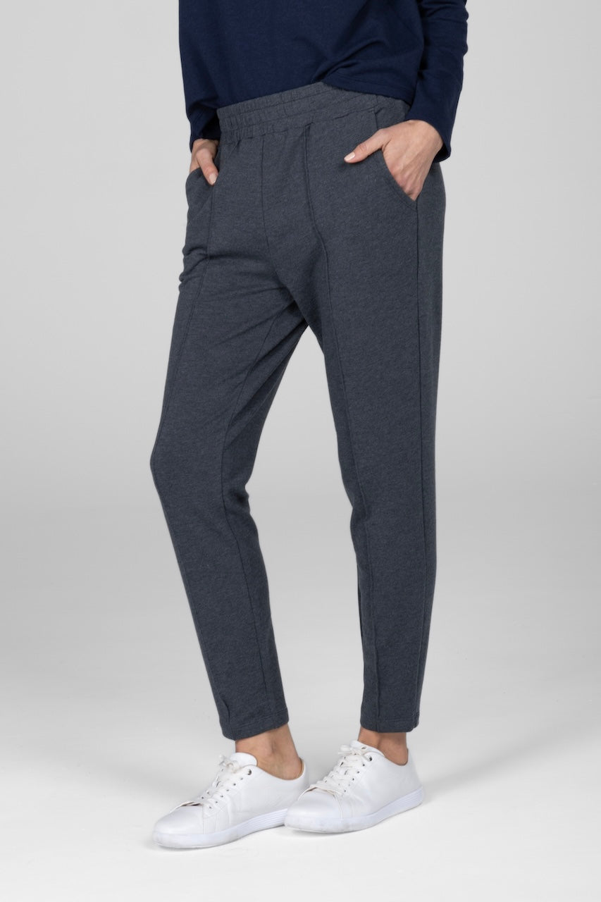 A woman standing, wearing charcoal heather travel pants with a checkered pattern on the leg, paired with a navy blue sweater and white sneakers.