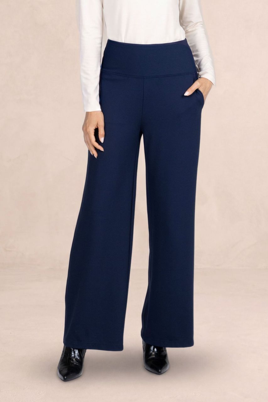 A model wearing navy blue wide-leg trousers with a high waistband, styled with a white long-sleeve top and black patent leather shoes, standing against a neutral beige backdrop.
