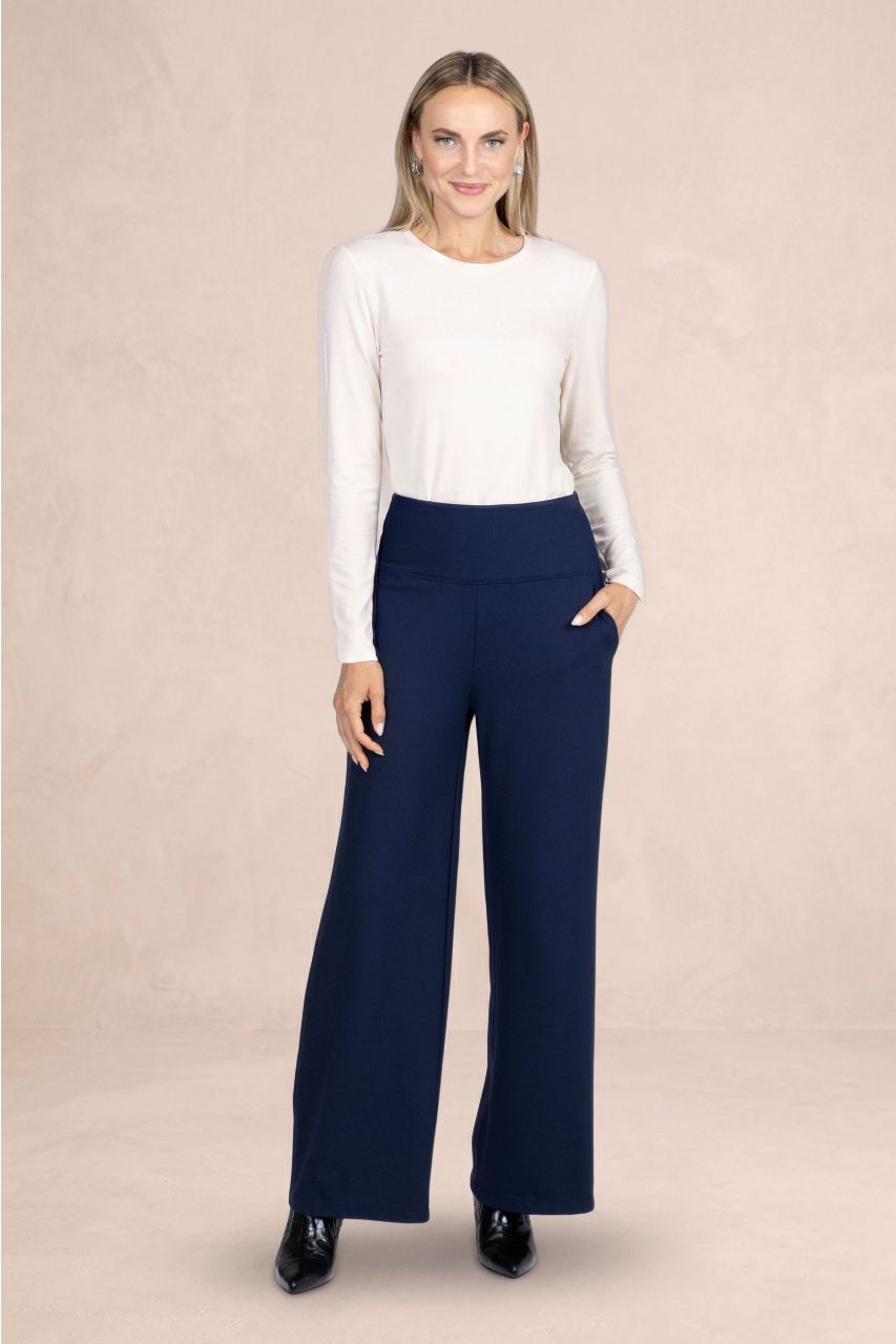 A woman modeling a white long-sleeve crewneck top and navy blue wide-leg trousers against a textured beige studio backdrop.