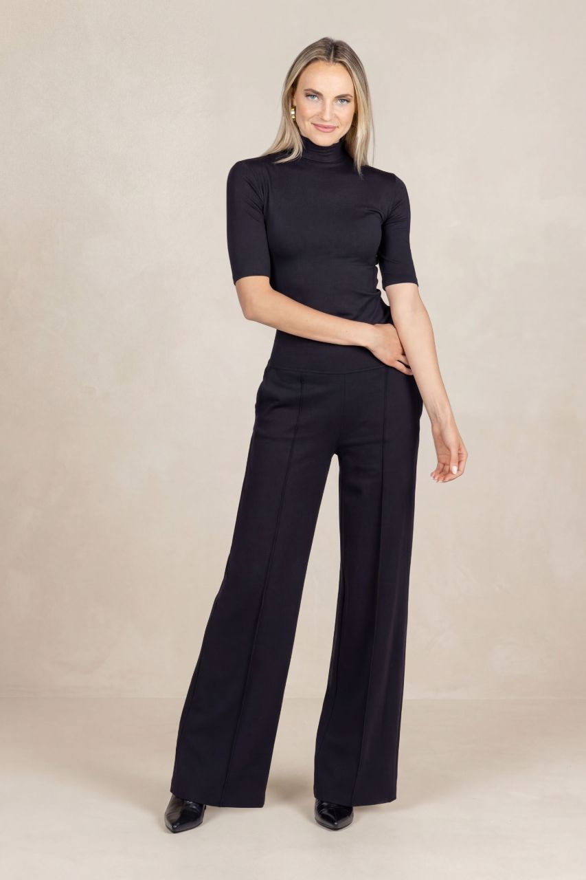 Woman wearing a black short sleeve mock neck and black wide-leg pants on a beige background
