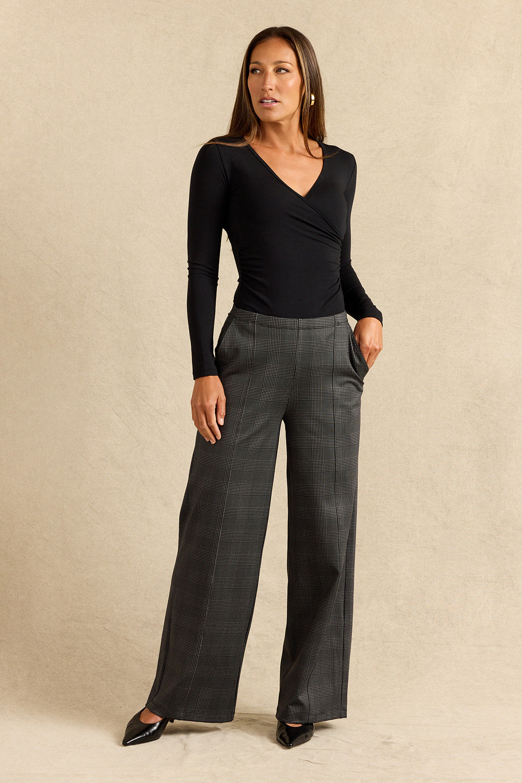 A woman modeling a black long-sleeve wrap-style top and dark charcoal gray wide-leg plaid trousers, standing against a textured beige studio backdrop.