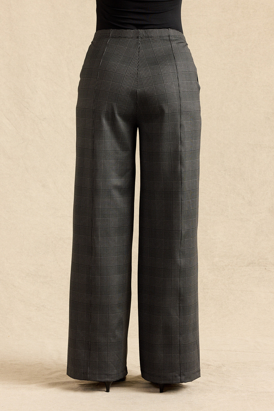 A pair of dark gray wide-leg plaid trousers shown from the back, worn by a model with a black top and heels, against a textured beige studio backdrop.