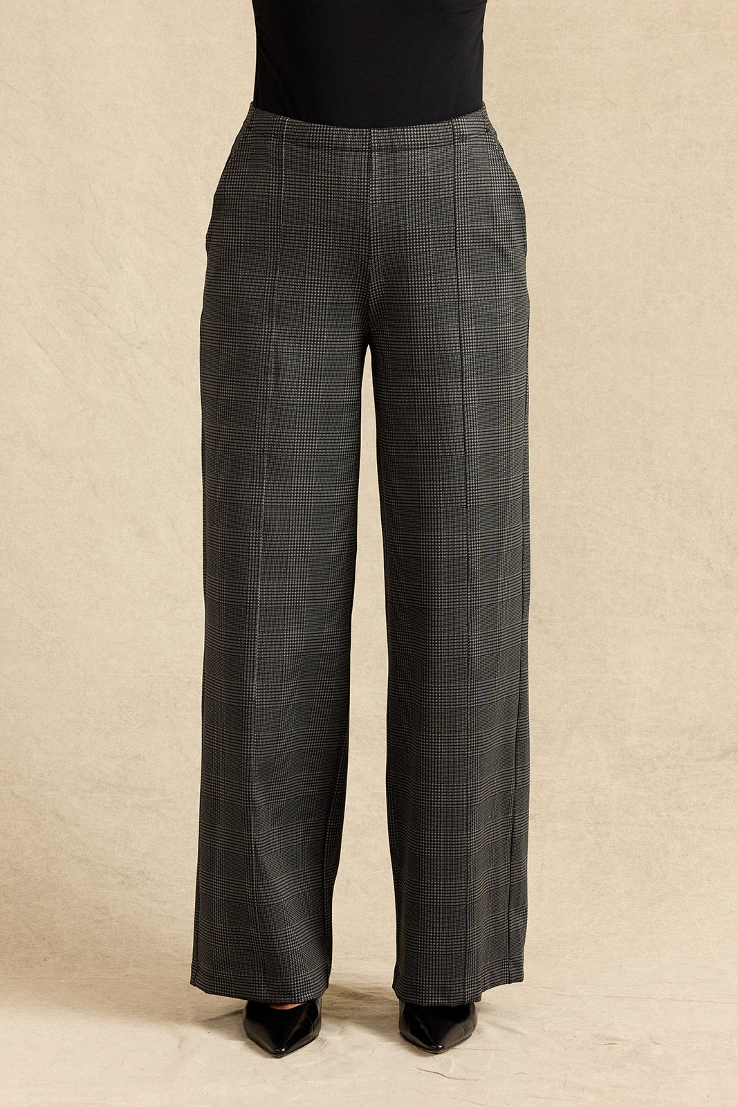 A person wearing dark charcoal gray wide-leg plaid trousers with a subtle windowpane pattern, paired with a black top and black pointed-toe heels, standing against a textured beige backdrop.