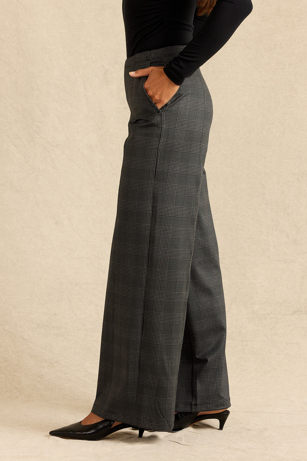 A person wearing high-waisted, wide-leg gray plaid trousers with a black long-sleeve top and black pointed-toe heels, standing against a textured beige backdrop.