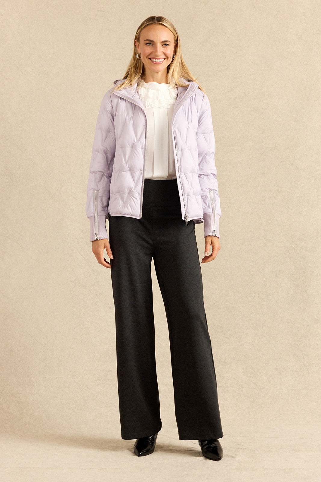 A woman modeling a light lavender hooded puffer jacket over a white ruffled top and charcoal wide-leg trousers, standing against a neutral beige studio backdrop.