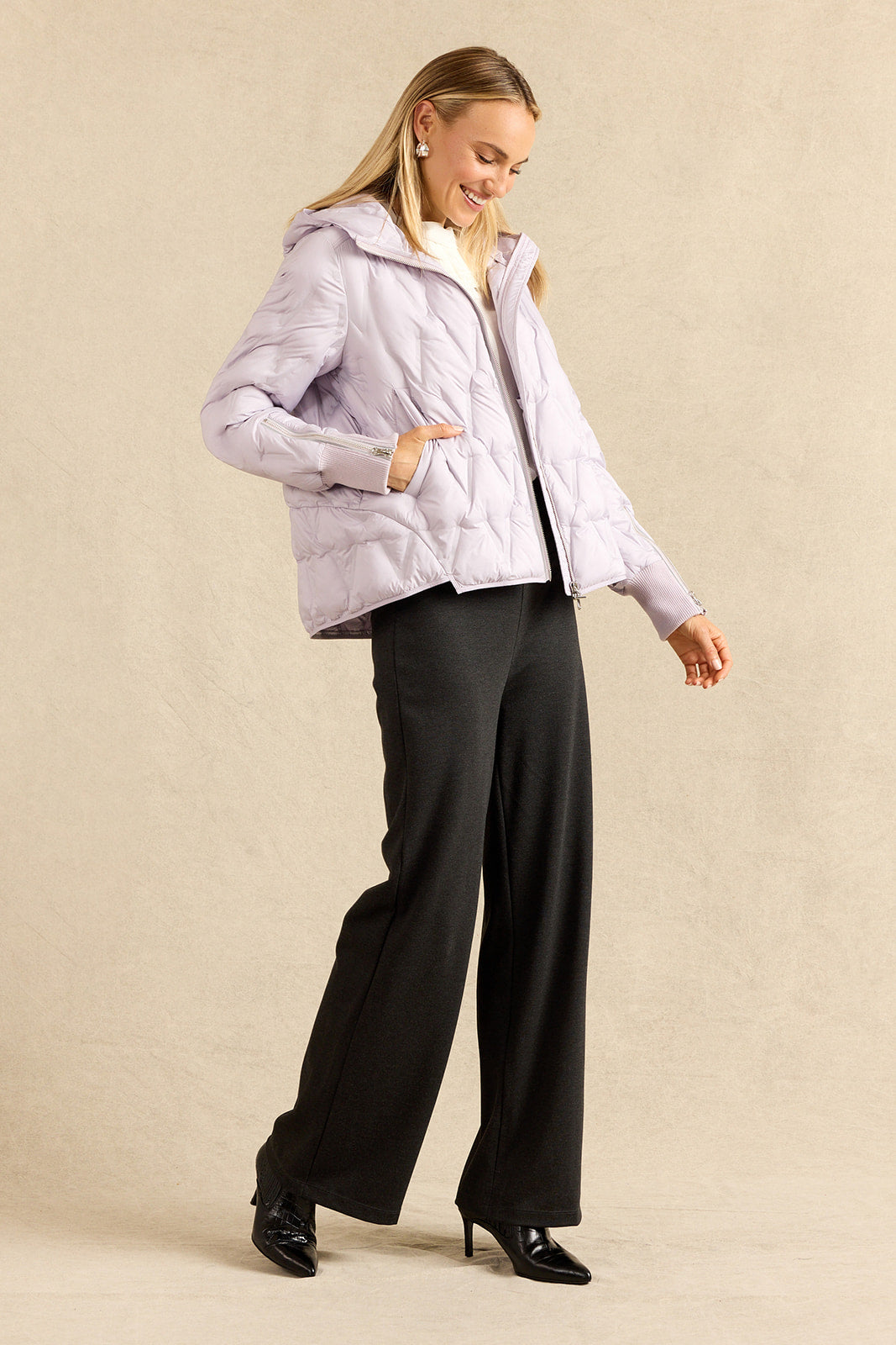 A woman modeling a light lavender quilted puffer jacket with a white blouse, paired with charcoal wide-leg trousers and black heels, against a neutral beige studio backdrop.