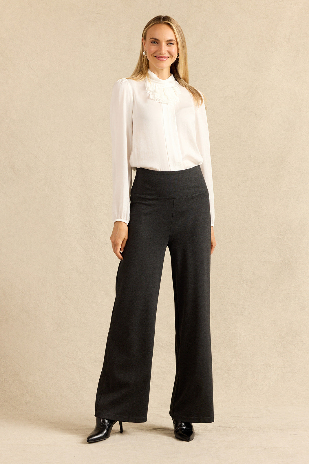 A woman modeling a white long-sleeve blouse with a ruffled high neck and charcoal wide-leg trousers, standing against a textured beige studio backdrop.