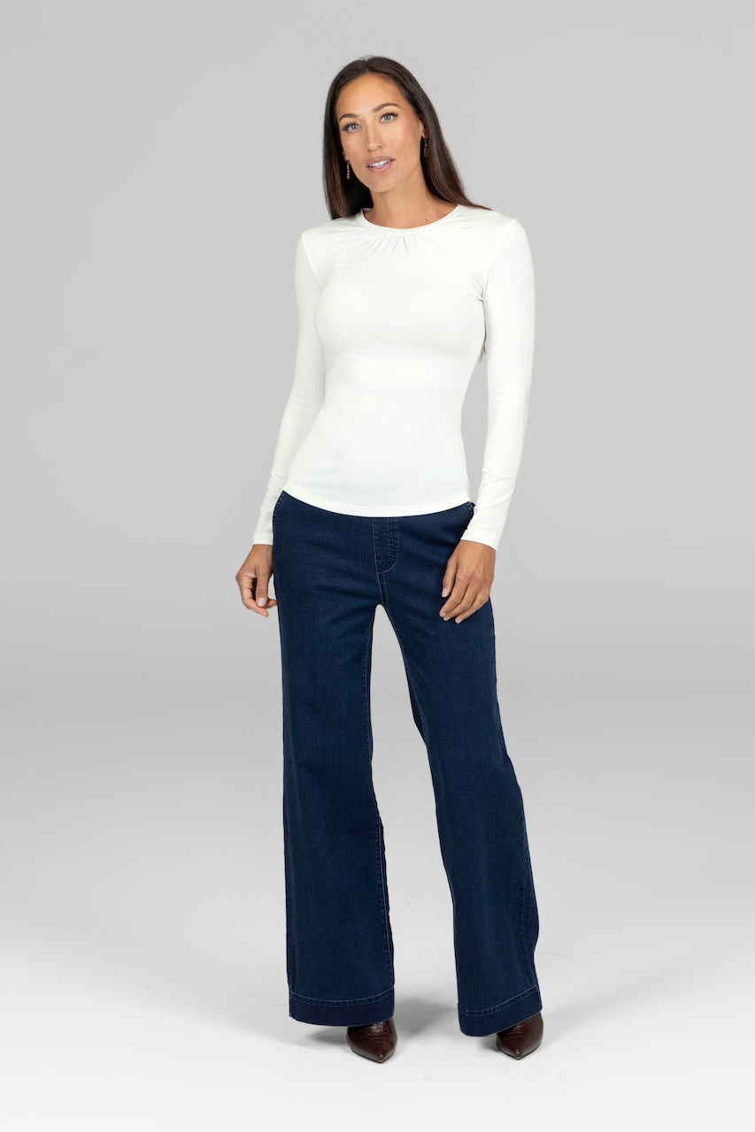A woman standing wearing a white long-sleeve top and dark wash stretch denim trousers.