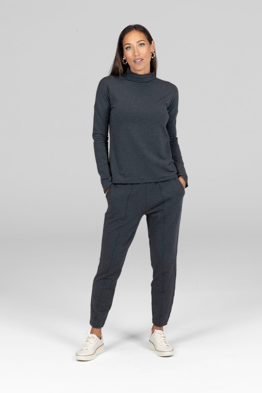 A female model wearing a charcoal heather gray mock neck pullover and matching pants, standing against a plain background.