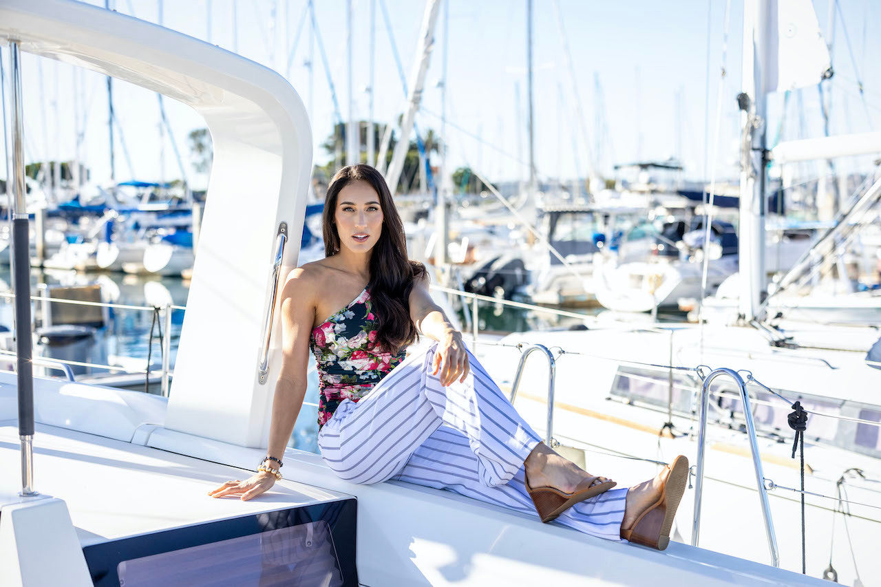 Model sitting on a boat in GODDESS LUXE ONE SHOULDER: BRIGHT SPRING FLORAL and stripped pants 