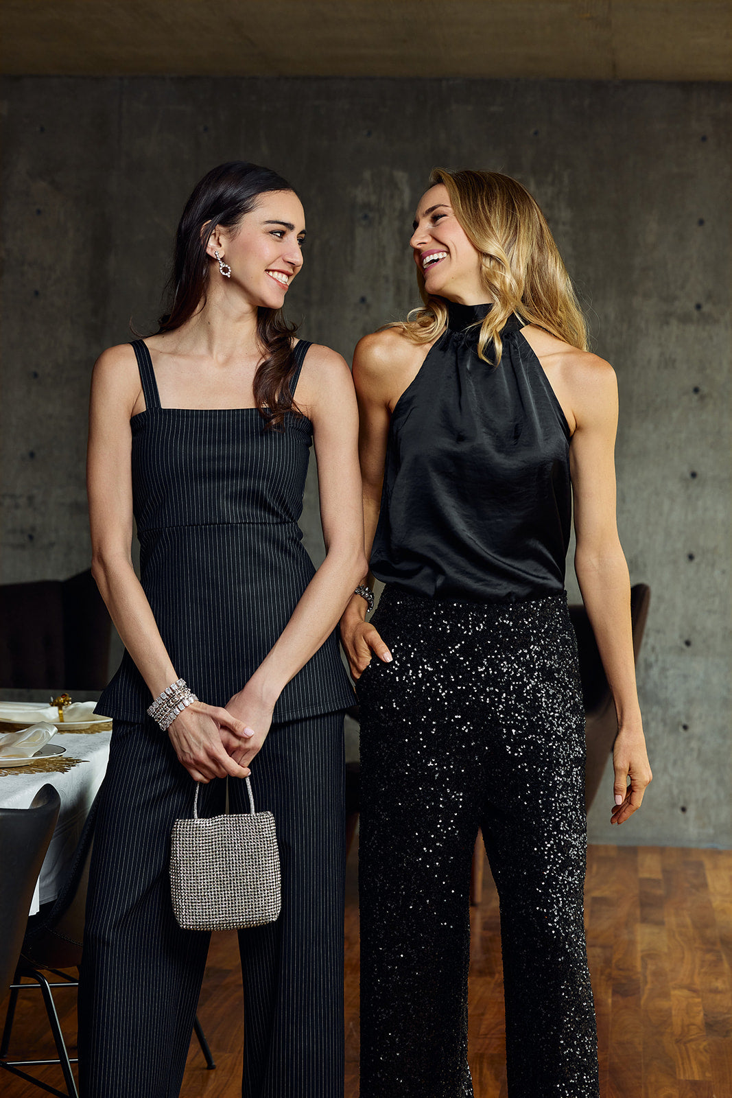 Two women standing indoors against a textured gray wall, wearing black outfits. The woman on the left wears a black pinstripe sleeveless top and matching wide-leg pants with a small gray handbag.