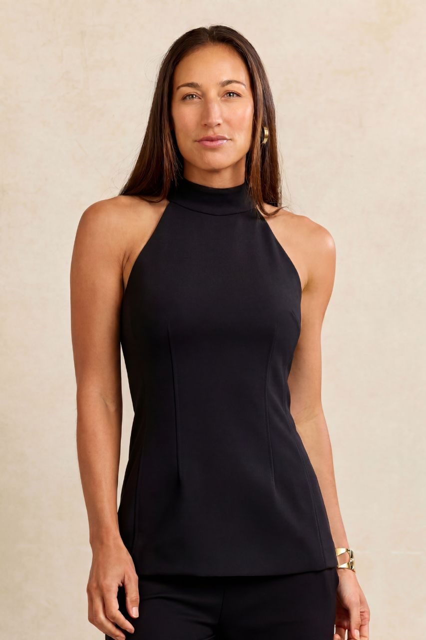 A woman modeling a sleeveless black mock neck top with structured side seams, paired with black pants, against a textured beige background.