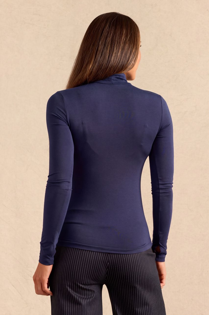 A woman shown from the back wearing a fitted navy blue long-sleeve turtleneck top and dark pinstriped pants, standing against a textured beige wall.