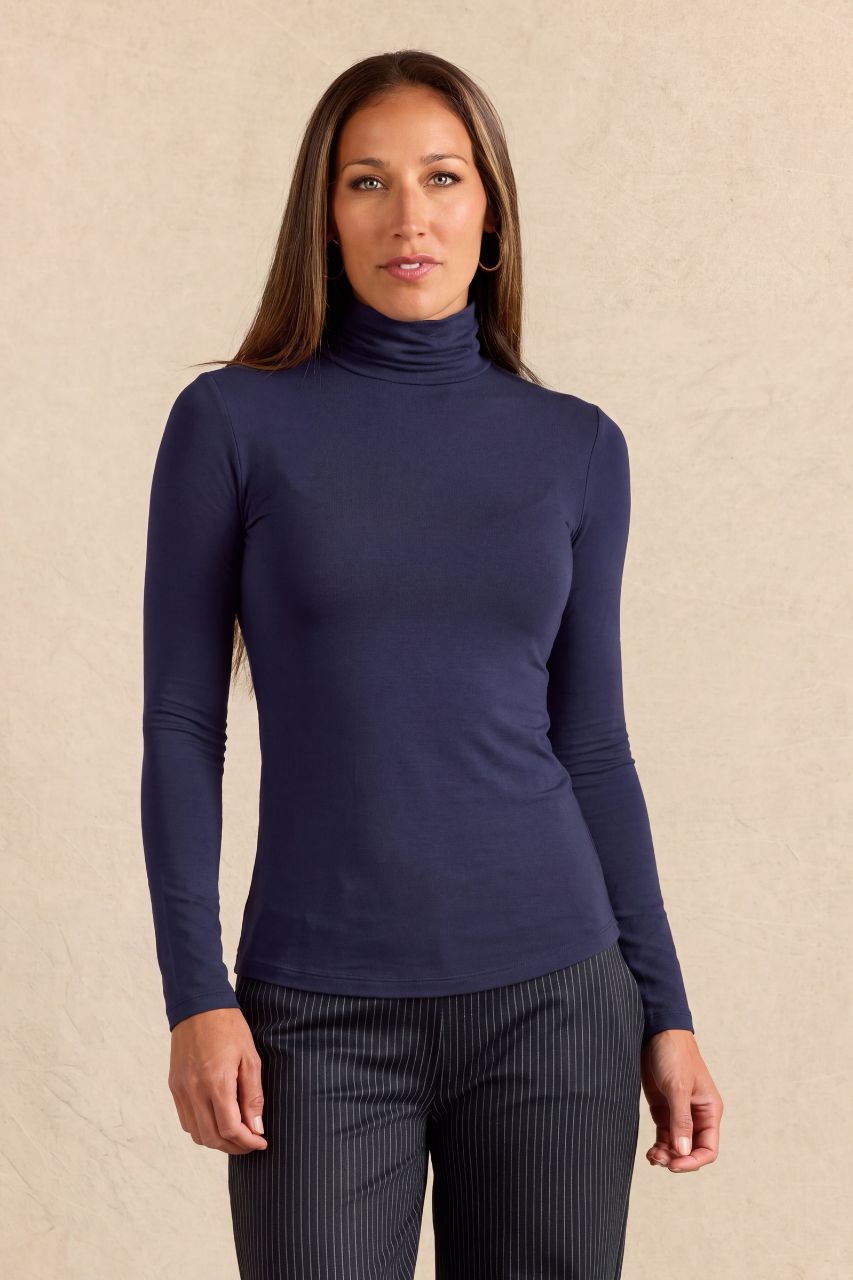 A woman modeling a navy blue long-sleeve turtleneck top against a textured beige background.
