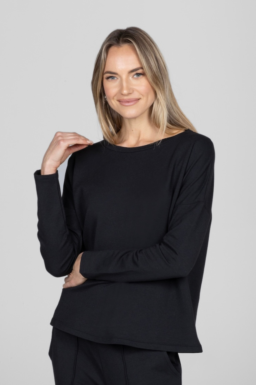 A woman wearing a black long sleeve top with a boat neckline and one hand on her shoulder.