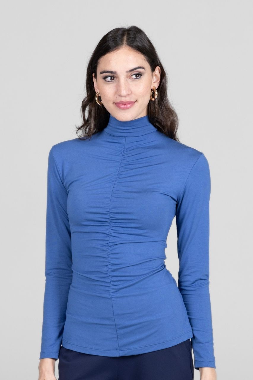Woman wearing a blue long-sleeve mock neck top with ruching down the front against a gray background