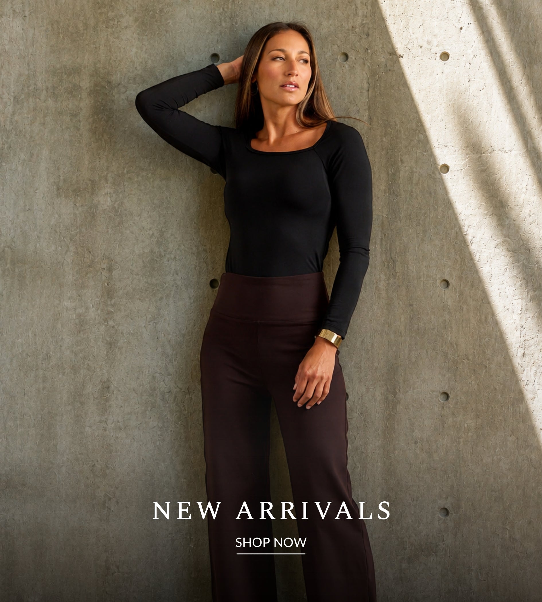 A woman posing against a textured concrete wall wearing a black long-sleeve square-neck top and dark brown wide-leg trousers. Text overlay at the bottom reads 'NEW ARRIVALS SHOP NOW'.