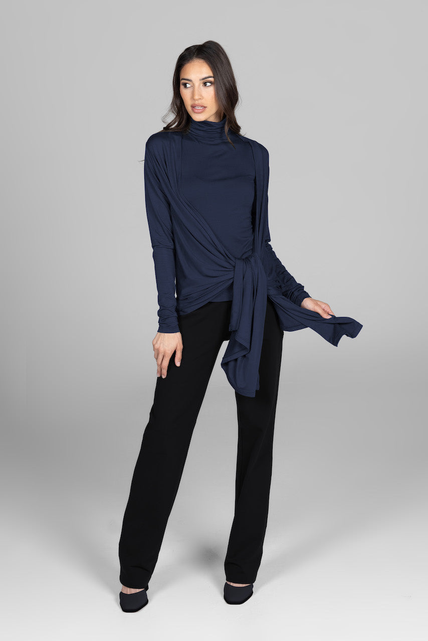 A woman wearing a long-sleeve navy blue wrap cardigan tied in the front over a matching turtleneck top, paired with black wide-leg pants and black shoes. She stands confidently, looking slightly to the side against a plain grey background.